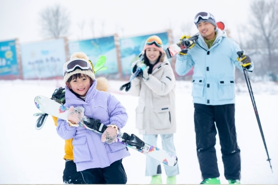 A young child holding skis, being watched by his parents
